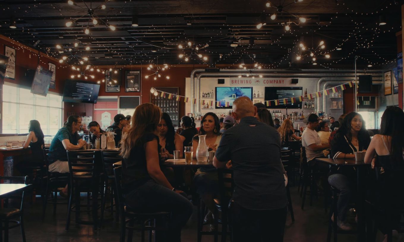 Customers inside of a brewery's restaurant sit at tables and have conversations, over food and drinks.