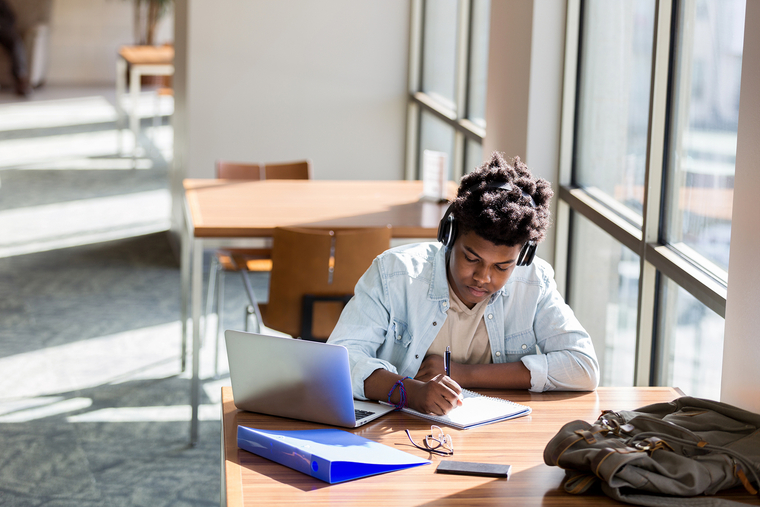 Young university student, wearing wireless headphones, writes in notebook while focused deeply on studying in bright, modern library